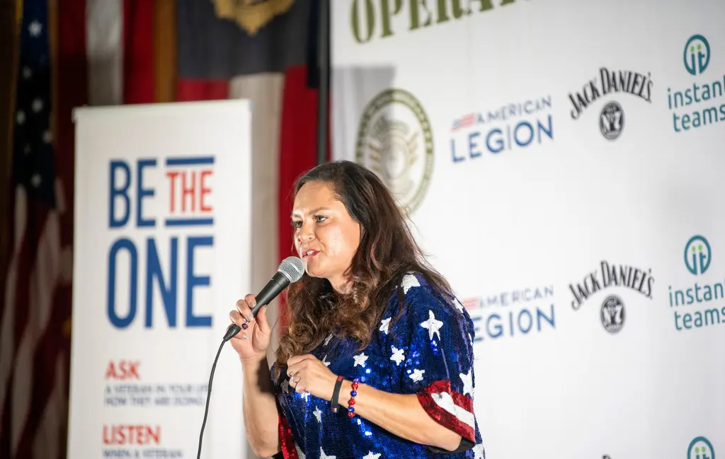 Retired Lt. Col. Robin “Phoenix” Johnson, in a sequined stars-and-stripes top, speaks into a microphone beside a Be the One banner.