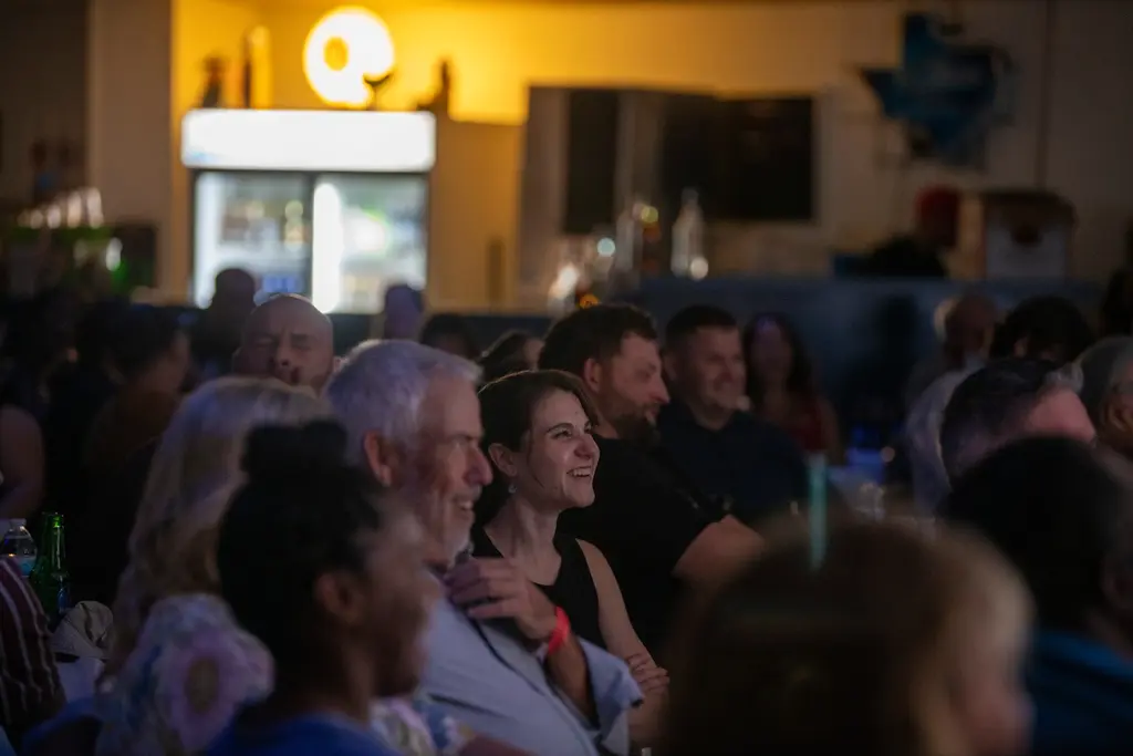 A crowded, dim hall of seated guests laughs toward the stage, with a smiling woman in a black top at center and a lit drinks cooler in the background.