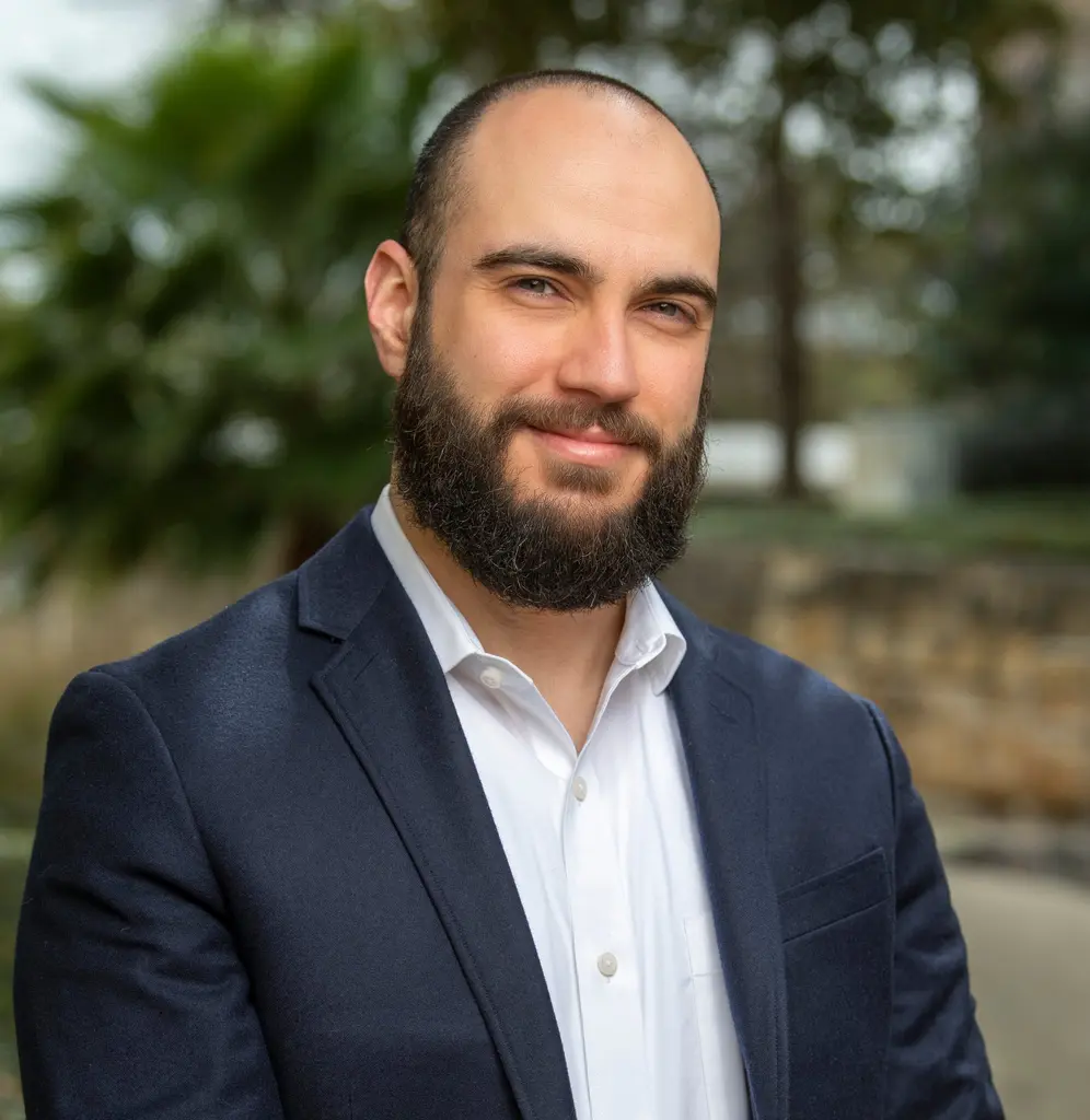 A bearded man in a navy blazer and white shirt smiles outdoors, with palms and a stone wall blurred behind him.