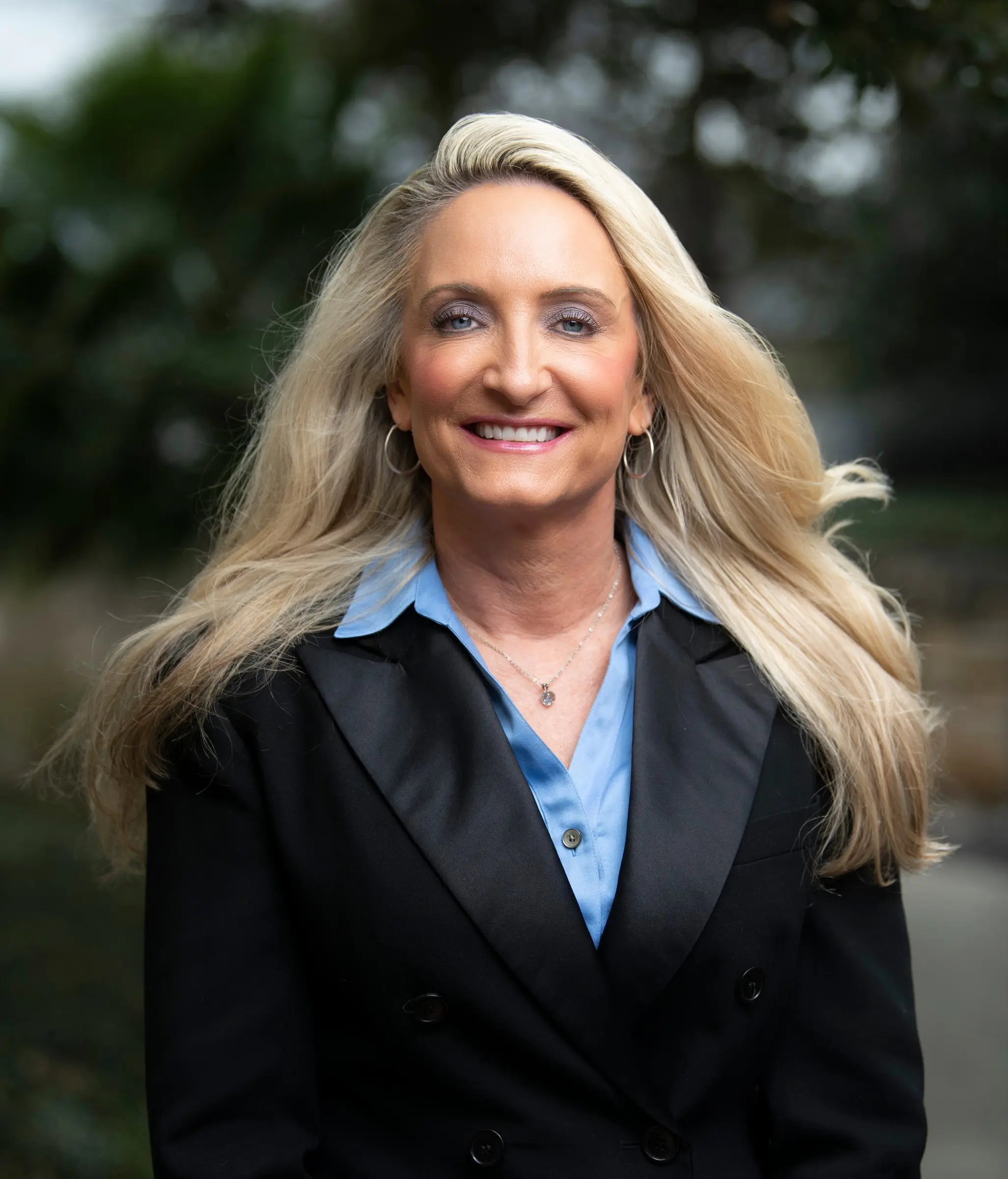 A woman with long, windblown blonde hair, in a black double-breasted blazer and a blue shirt, smiling outdoors.