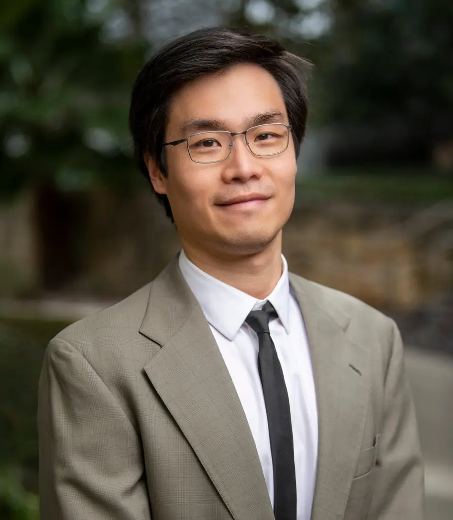 A man in glasses, a tan suit and a slim black tie, photographed outdoors against blurred trees and stone.