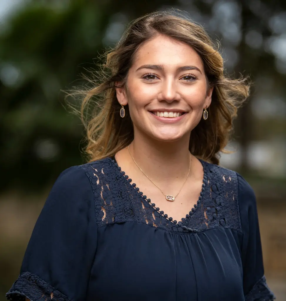 A young woman with wavy, windblown hair, in a navy lace-trimmed blouse, smiles broadly outdoors.