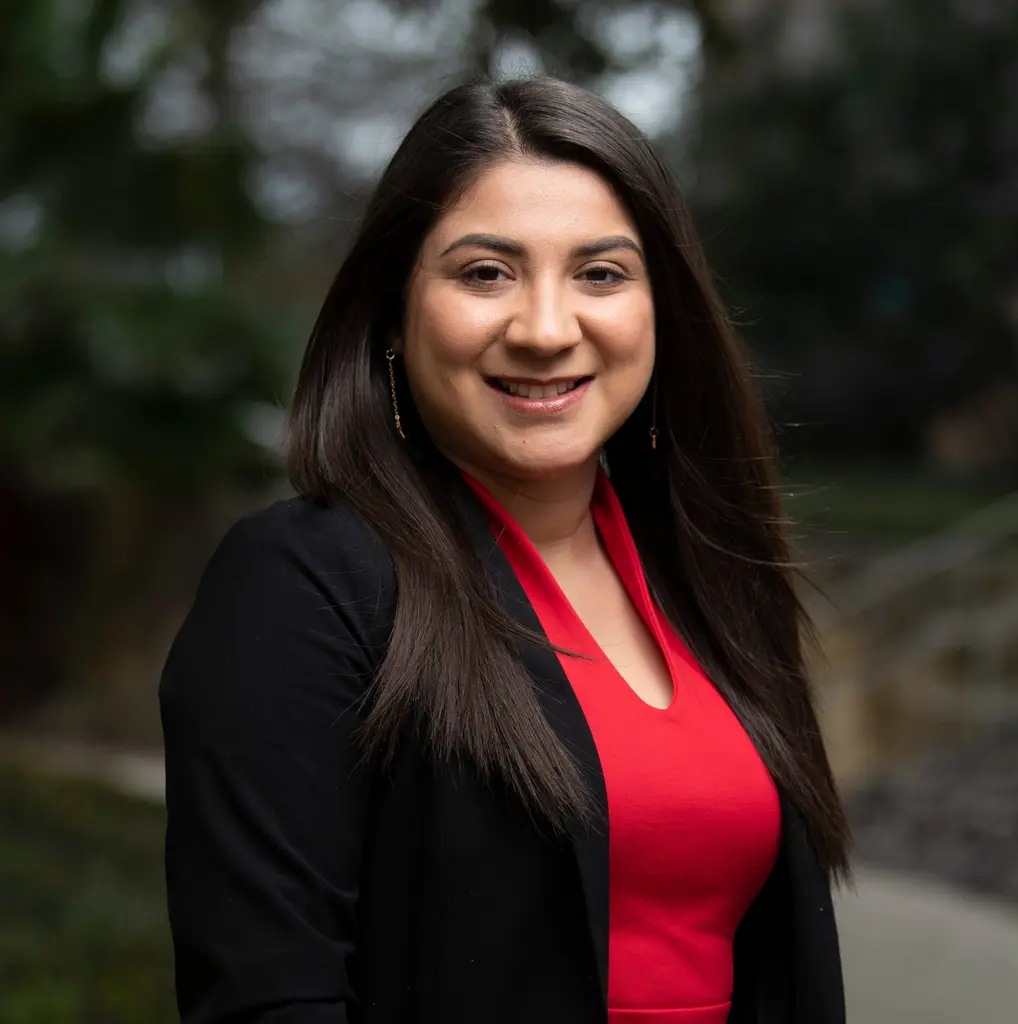 A smiling woman with long dark hair, in a red top and a black blazer, photographed outdoors.