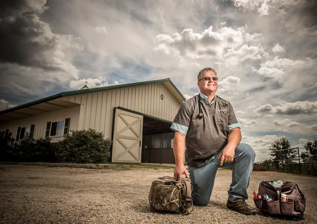 Dr. Patrick O'Neil kneels between two veterinary bags in front of a barn under a cloudy sky
