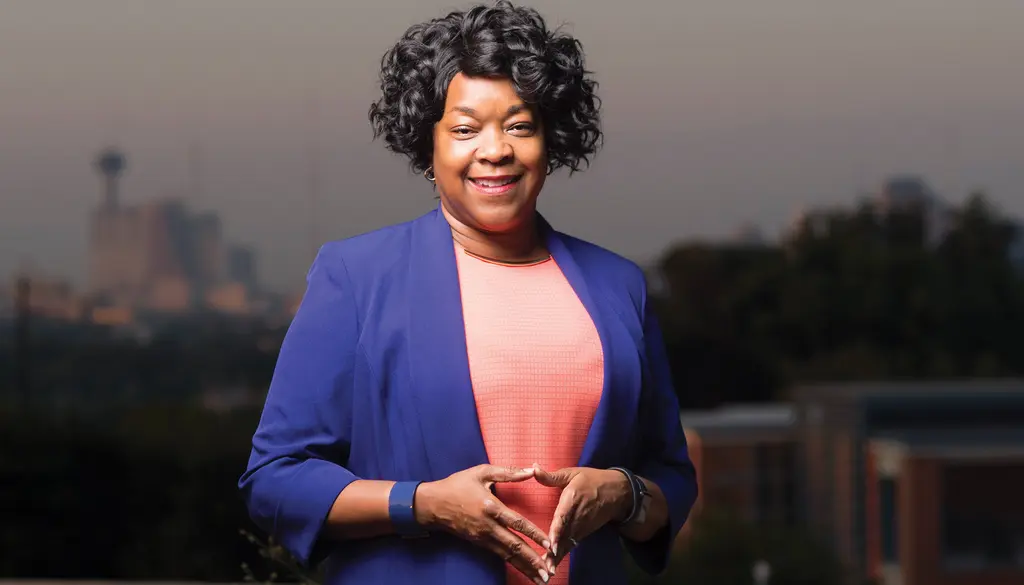 Paula Gold-Williams in a royal blue jacket smiles with the Tower of the Americas and downtown skyline behind her