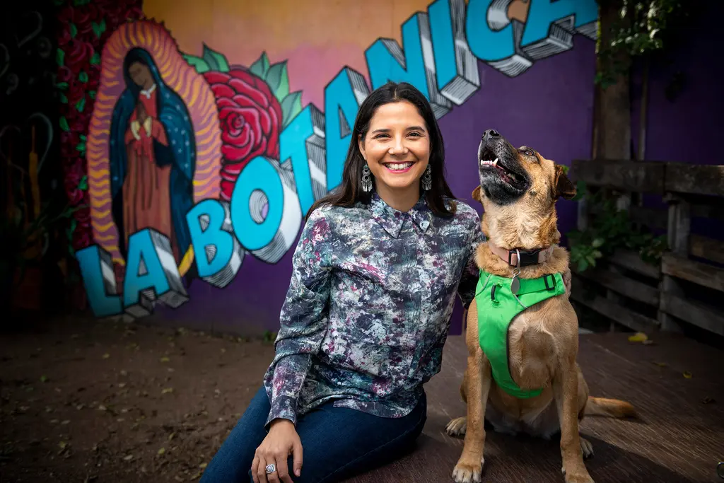 A smiling woman sits beside her dog in a green harness in front of a La Botanica mural of the Virgin of Guadalupe and a red rose.