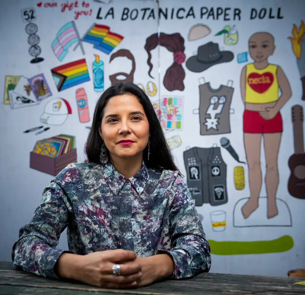Rebel Mariposa sits at a table in front of a painted La Botanica paper doll mural