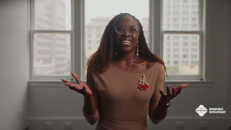 A presenter smiles with open hands in an office with tall windows, in the how-to video on starting a registered apprenticeship program.
