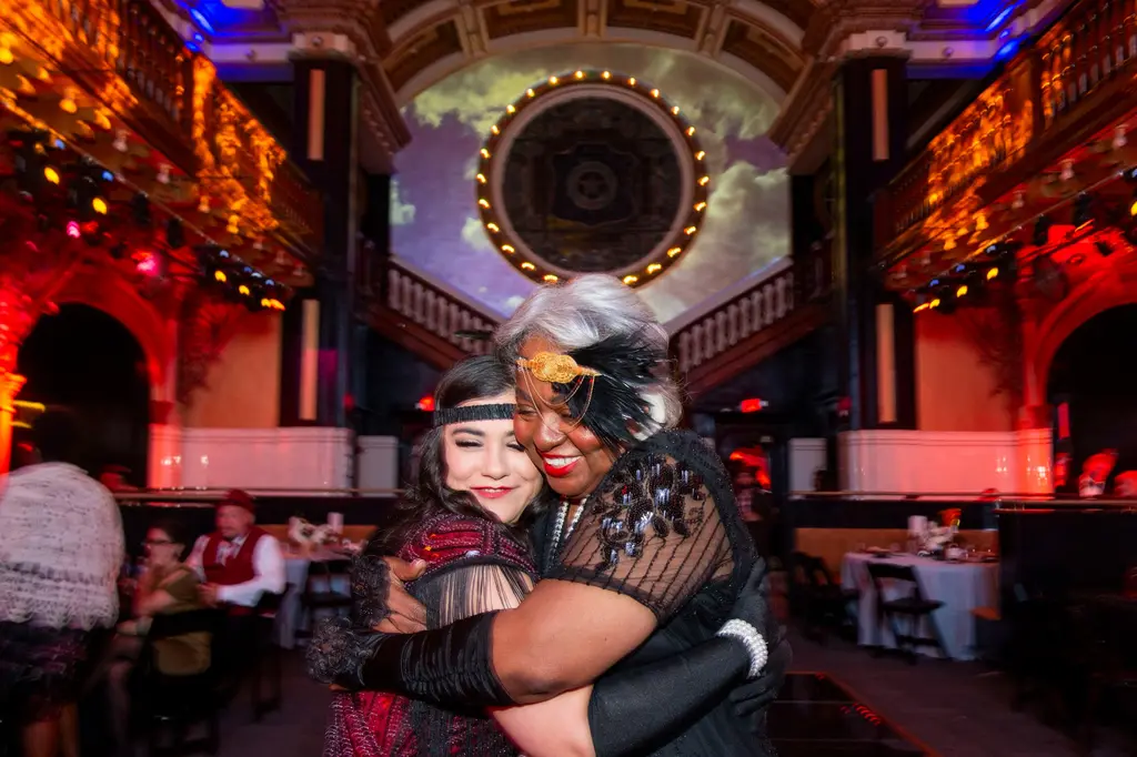 Two women in flapper headbands hug and smile in the middle of the hall, a round window lit up behind them.