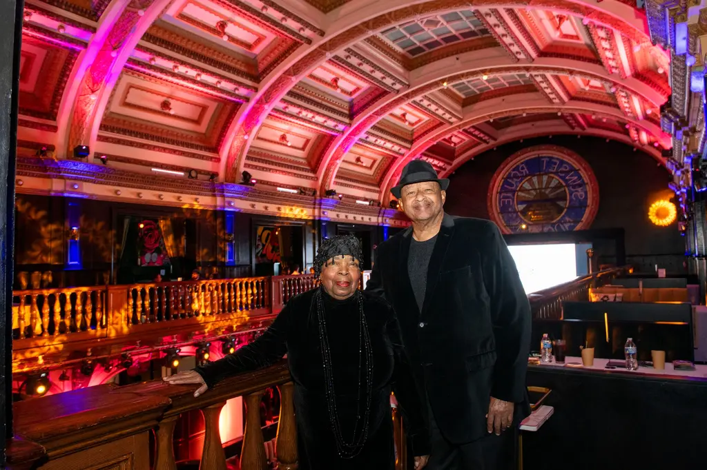 A woman in a black beaded outfit and a man in a black velvet jacket and hat stand at a wooden balcony rail in a hall with a pink-lit ceiling and round stained glass window.