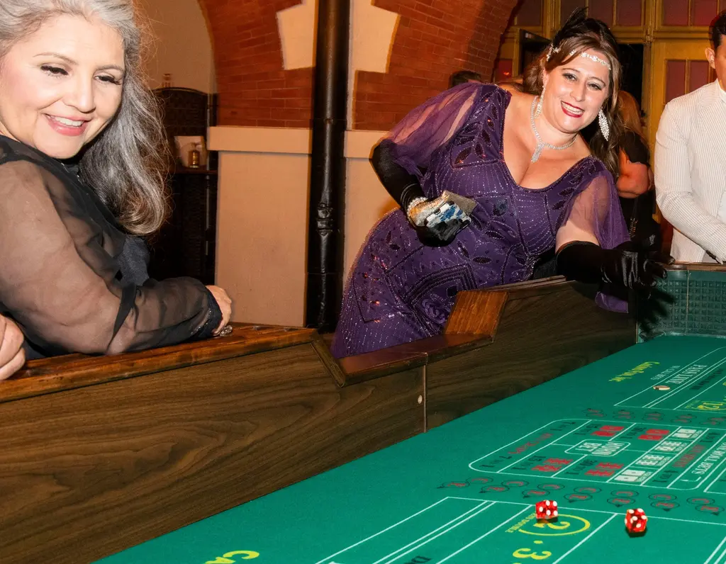A smiling woman in a purple beaded dress and black gloves throws dice down a green craps table as another woman watches.