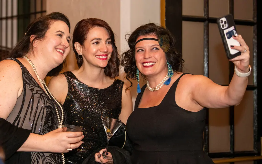 Three smiling women in black dresses and pearls lean together while one holds up a phone for a selfie.