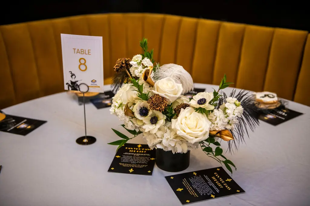 A round table with a white tablecloth holds a centerpiece of white roses, anemones and black feathers, a Table 8 card and printed cards.