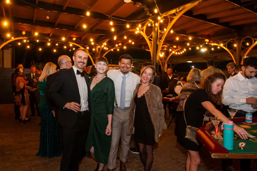 Guests in suits and dresses stand and smile around casino tables under iron arches strung with lights at night.