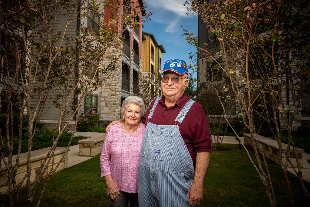 An older couple stands between young trees in an apartment courtyard, the man in overalls and a blue Shrine Circus cap.