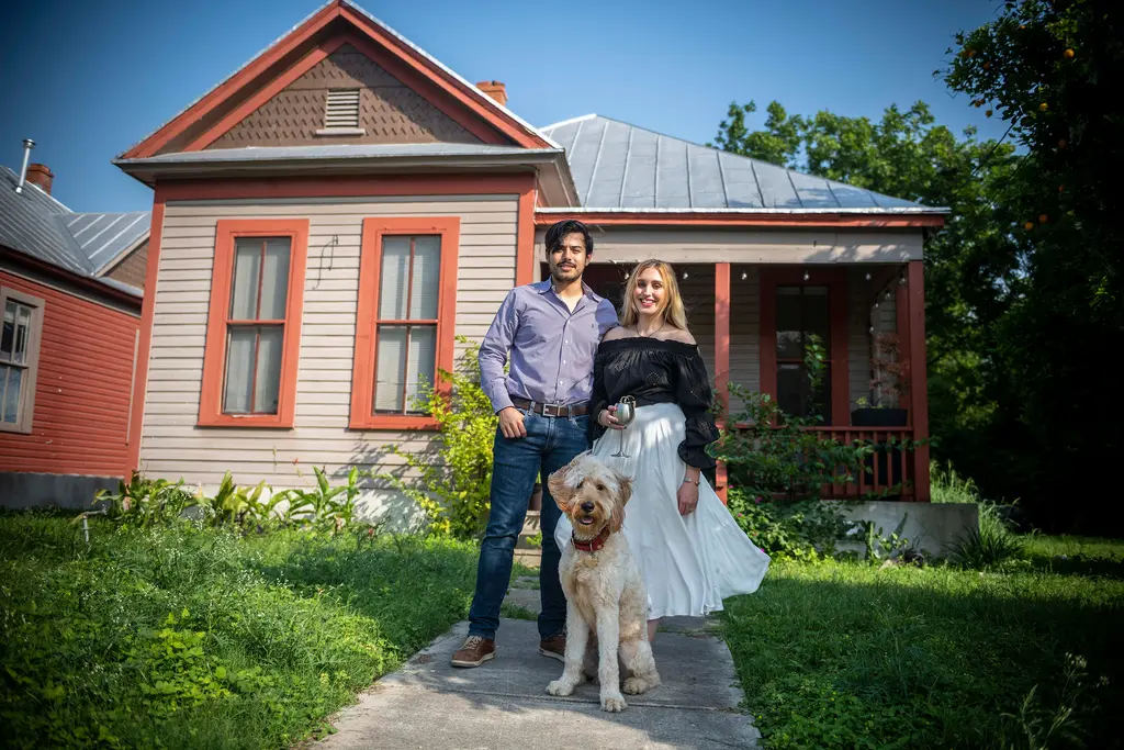 A couple stands on the front walk of a gray and red bungalow with their shaggy golden dog sitting in front of them.