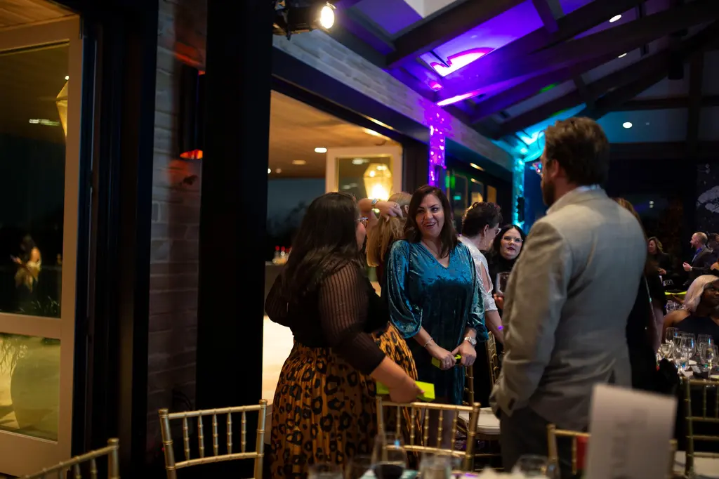 A woman in a teal velvet dress smiles while talking with a woman in a sheer black top and a man in a tan blazer, beside open glass doors under purple and blue uplighting.