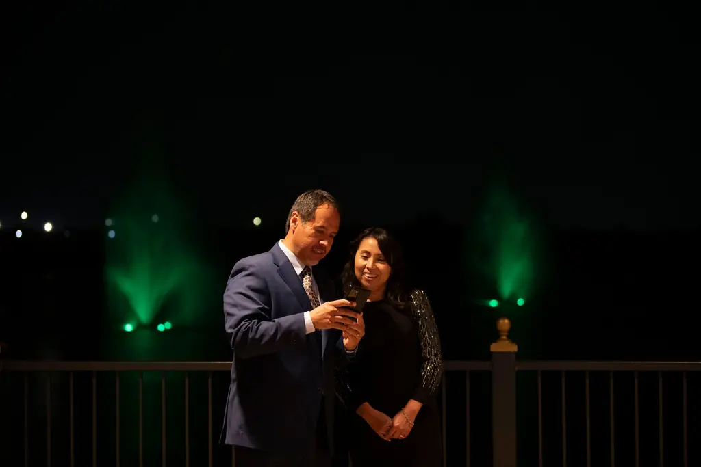 A man in a navy suit and patterned tie and a smiling woman in a black dress with sparkling sleeves look at a phone screen at night, with green-lit fountains in the water behind them.