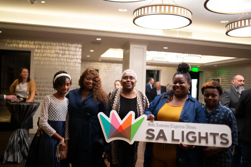 Five smiling guests, from a girl in a white headband to a woman in a denim jacket and a boy in a patterned shirt, stand behind a large handheld sign reading San Antonio Express-News SA Lights with a multicolored crown logo.