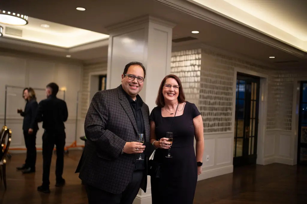 A smiling man in glasses and a checked jacket stands beside a smiling woman in a black dress, each holding a flute of sparkling wine, with white paneled walls behind them.