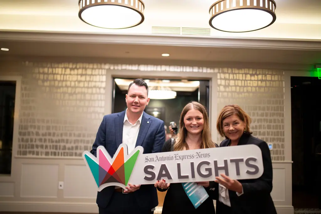 A man in a blue checked blazer and two smiling women, one with long light-brown hair and one in a dark jacket, hold the SA Lights sign in front of a doorway with a chandelier beyond.