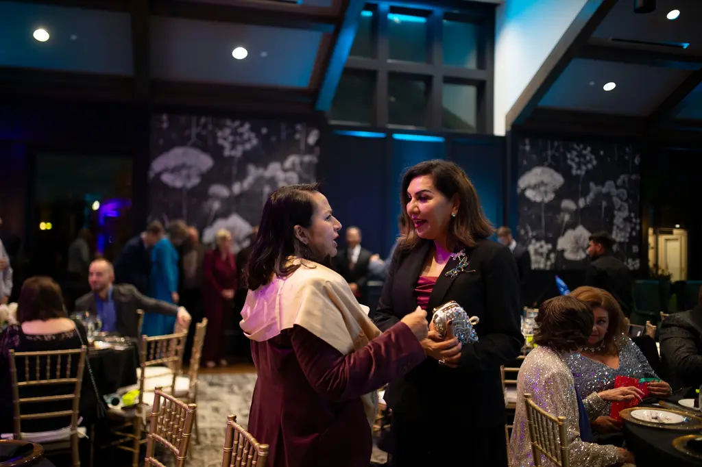 A woman in a cream shawl and a woman in a black jacket with a jeweled dragonfly brooch talk among gold chairs and set tables, with large white flower murals and blue uplighting behind them.