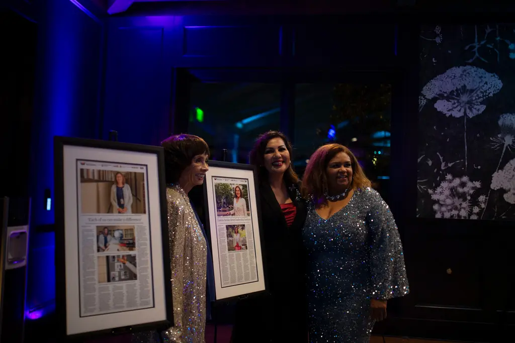 Three smiling women, one in a silver sequined jacket and one in a blue sequined gown, stand between framed newspaper pages with photos and headlines on easels, under blue uplighting.