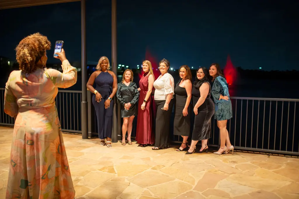 A woman in a long pastel floral dress raises her phone to photograph seven women in evening dresses posed along a railing, with red-lit fountains rising from the dark water behind them.