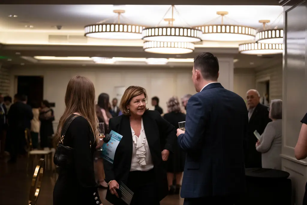 A woman with shoulder-length hair in a black blazer and ruffled white blouse smiles while talking with a man in a navy plaid suit and a woman holding a flute, under drum chandeliers.