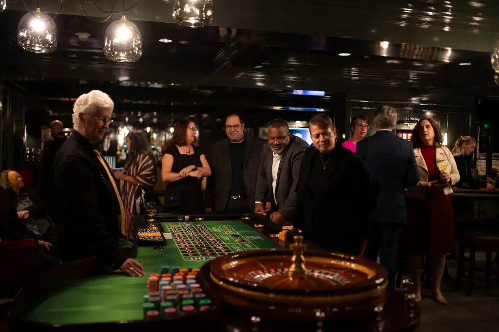 A white-haired dealer in a black shirt and gold tie stands at a roulette table with stacks of colored chips and a wooden wheel as guests in evening clothes look on under glass globe pendant lights.
