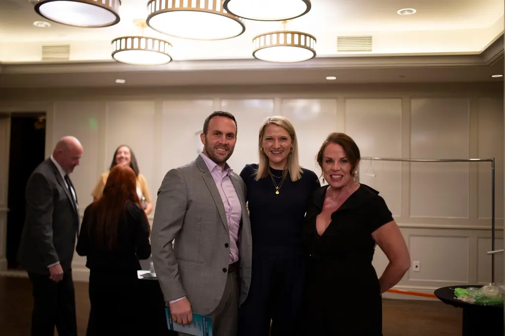 A man in a gray blazer and lavender shirt, a blond woman in navy and a woman in a black ruffled dress stand close and smile in a bright paneled room.