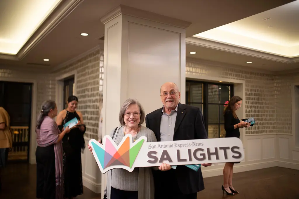 An older woman with gray hair and glasses and a bald man with glasses in a dark blazer hold the SA Lights sign in front of a white column and silver brushstroke wallpaper.