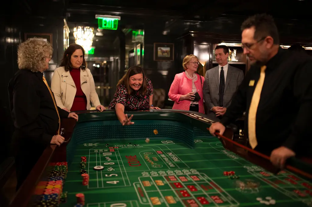 A laughing woman in a patterned top leans over a green craps table with her arm outstretched after a throw, while a dealer in a gold tie, a woman in a cream jacket and a couple in pink and plaid look on.