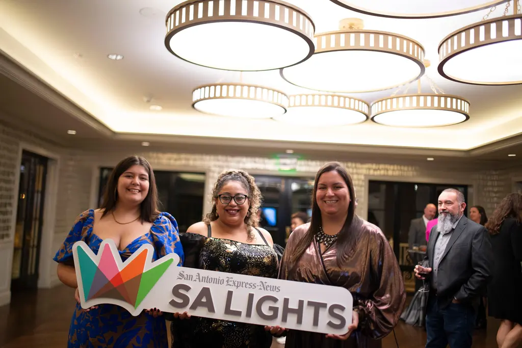 Three smiling women, in a blue floral gown, a black-and-gold sequined dress and a bronze satin dress, hold the SA Lights sign under round chandeliers.