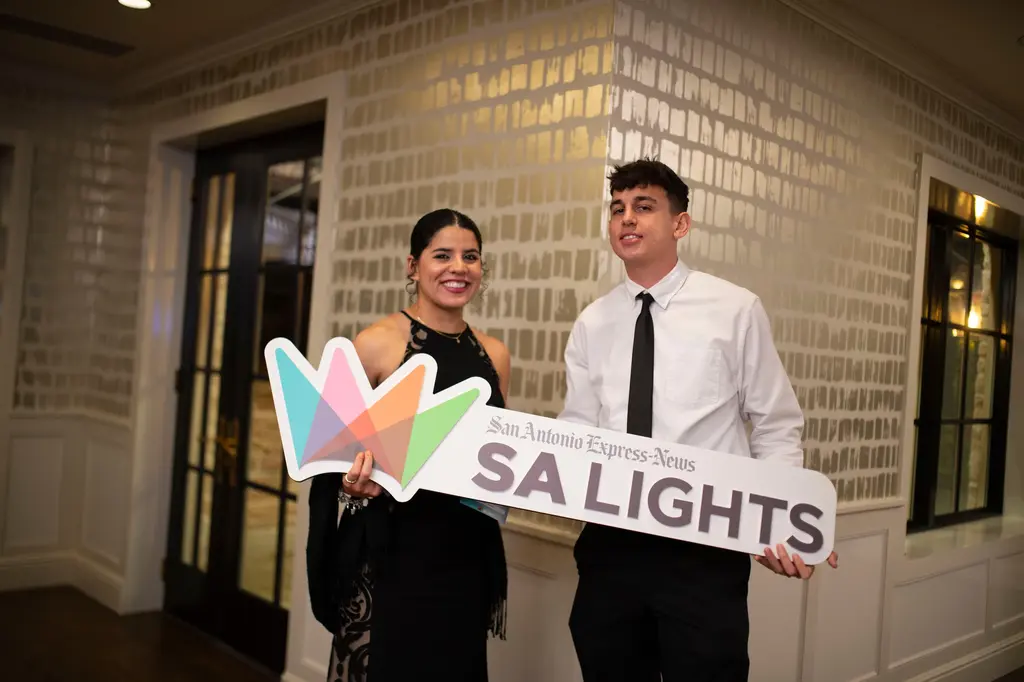 A smiling woman in a black halter top and a young man in a white shirt and black tie hold a tilted SA Lights sign with a colorful crown logo in front of silver brushstroke wallpaper.