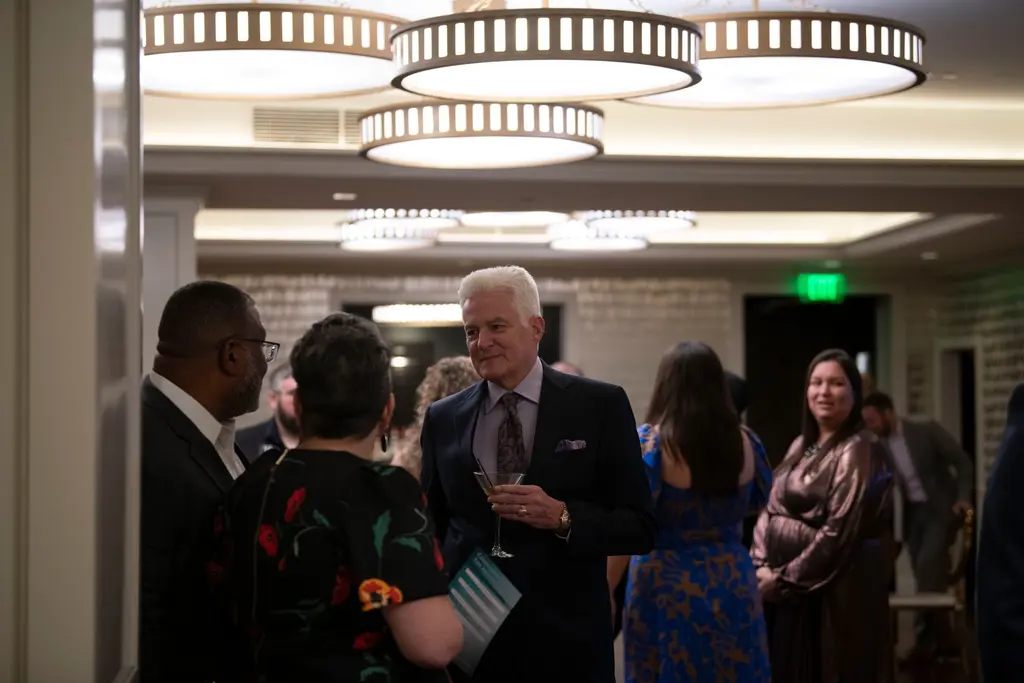 A silver-haired man in a dark suit holds a cocktail glass as he talks with guests under rows of drum-shaped chandeliers in a cream-paneled room.