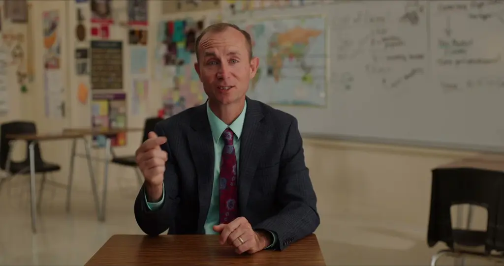 Clinton Roberson, in a dark plaid suit, a mint-green shirt and a red patterned tie, gestures as he talks at a desk in a classroom, with a whiteboard and a world map behind him.