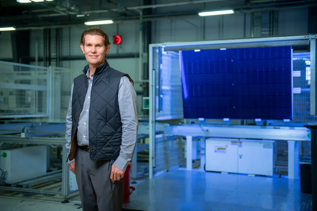 Sam Martens in a quilted navy vest stands on the factory floor beside a blue-lit solar panel on the production line.