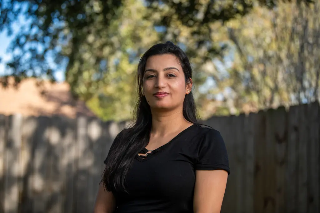 Anju Kumari, in a black top, stands in front of a wooden fence and trees.