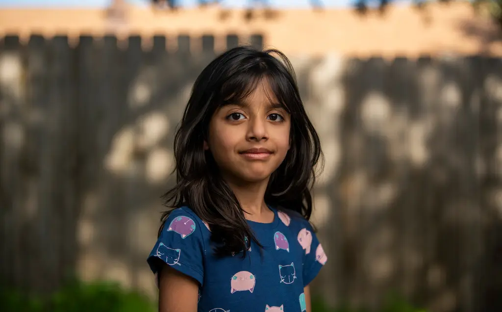 Anju Kumari’s daughter, in a blue print dress, stands in front of a wooden fence.