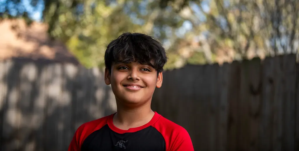 Anju Kumari’s son, in a red and black shirt, smiles in front of a wooden fence.