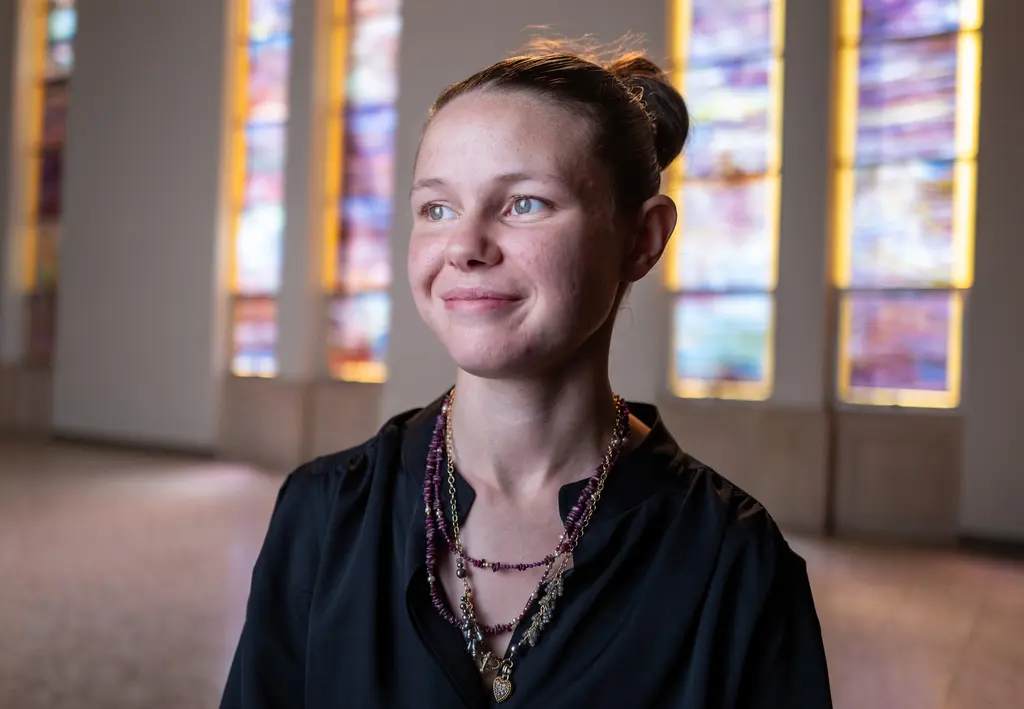 Ashley Keen, in a black top and beaded necklaces, smiles in a room with tall stained-glass windows.