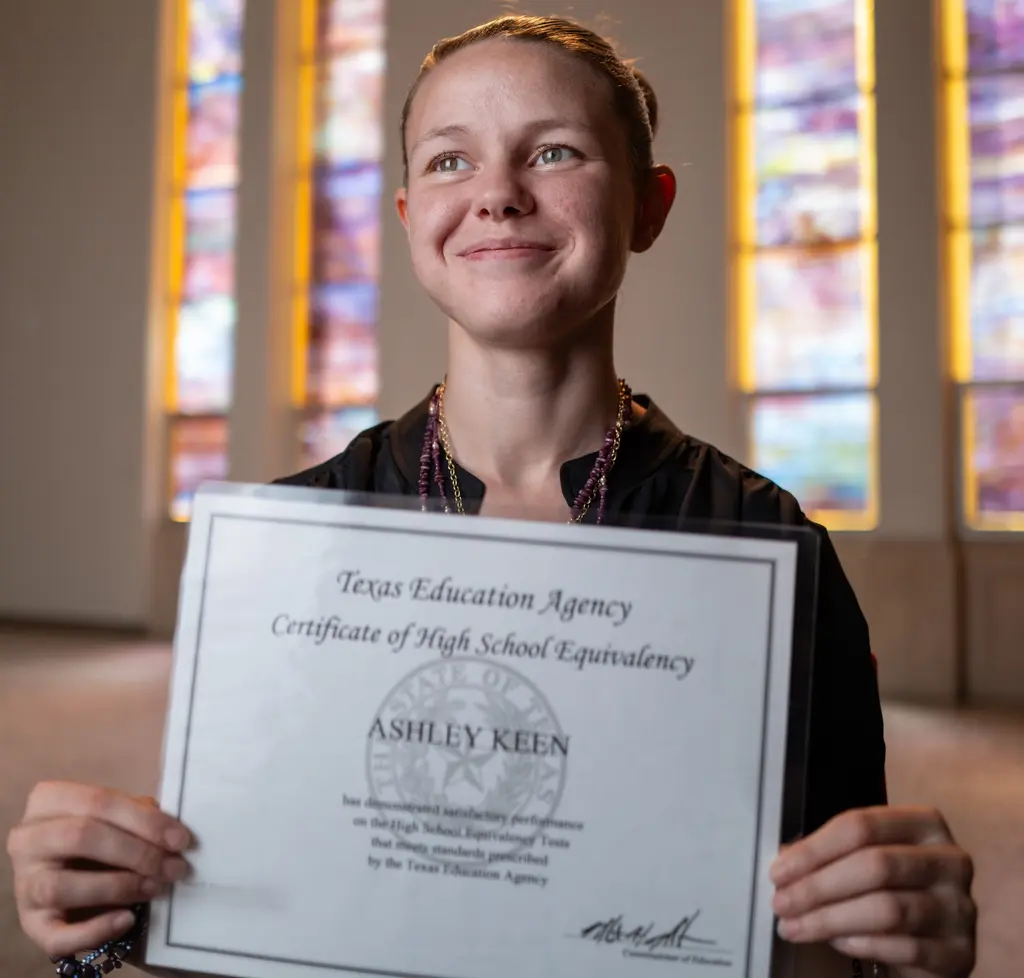 Ashley Keen holds her Texas Education Agency high school equivalency certificate in front of stained-glass windows.