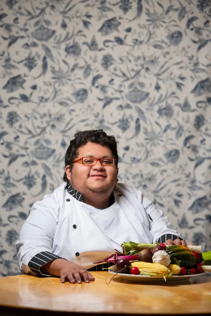 Jacob Gutierrez, in a white chef’s coat, sits at a wooden table with a platter of vegetables in front of floral wallpaper.