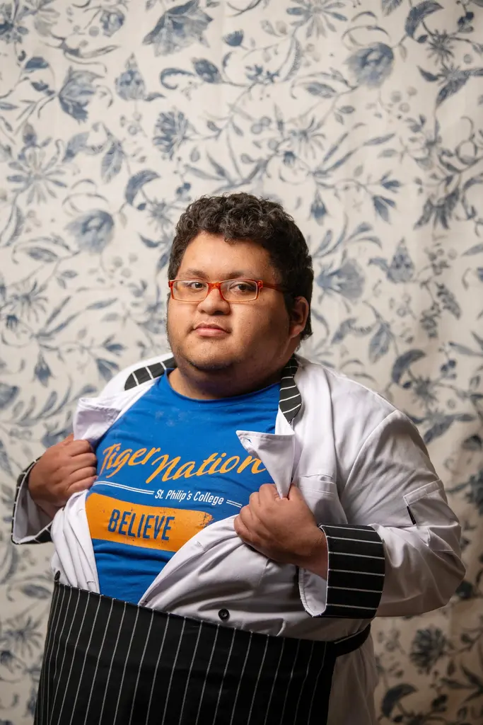 Jacob Gutierrez opens his white chef’s coat to show a blue St. Philip’s College “Tiger Nation” shirt in front of a floral backdrop.