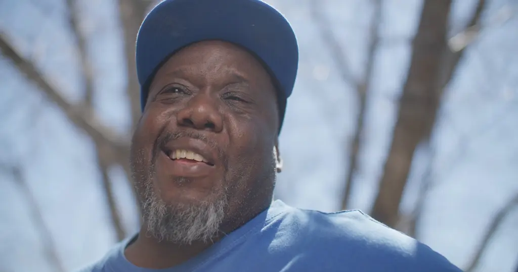 Scott Washington, in a blue cap and blue T-shirt, laughs under bare tree branches.