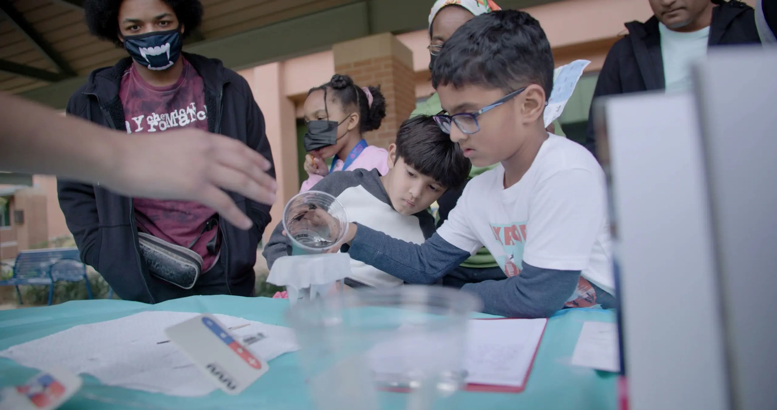 A boy in glasses tips a clear plastic cup at a demo table as other children and adults lean in to watch.