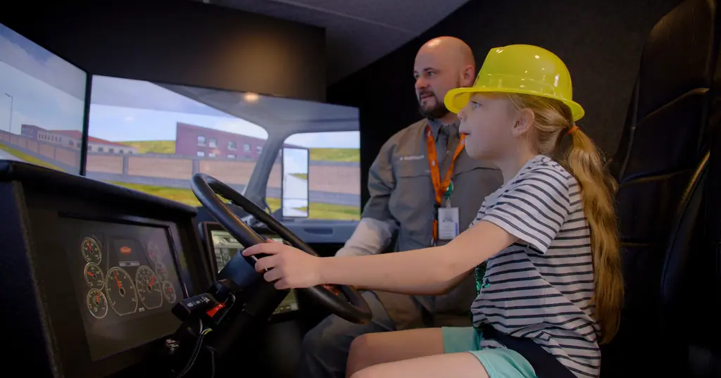 A girl in a yellow hard hat and striped shirt steers a truck-driving simulator as a man beside her, with an ID badge on an orange lanyard, watches the screens.