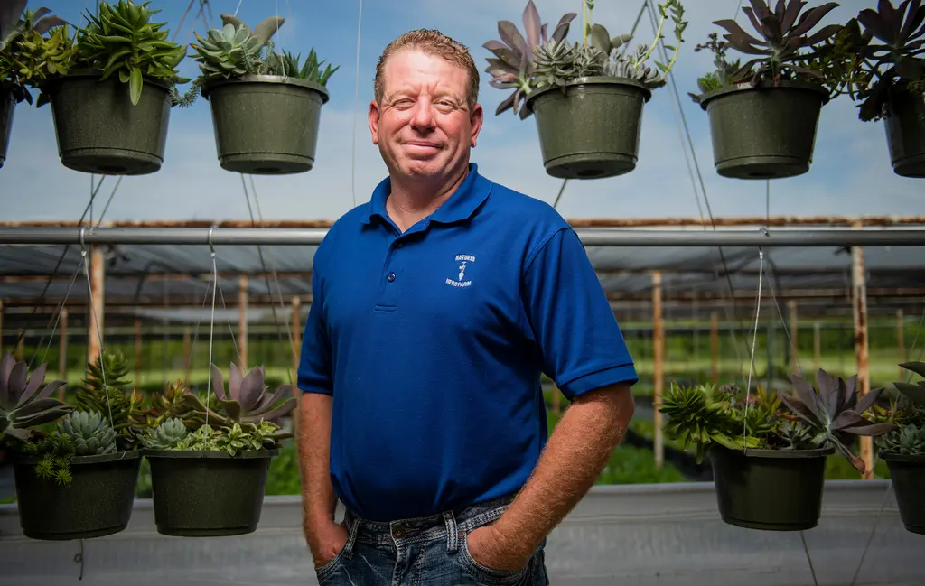 Shane Dunford in a blue polo stands beneath rows of hanging succulent baskets in a greenhouse