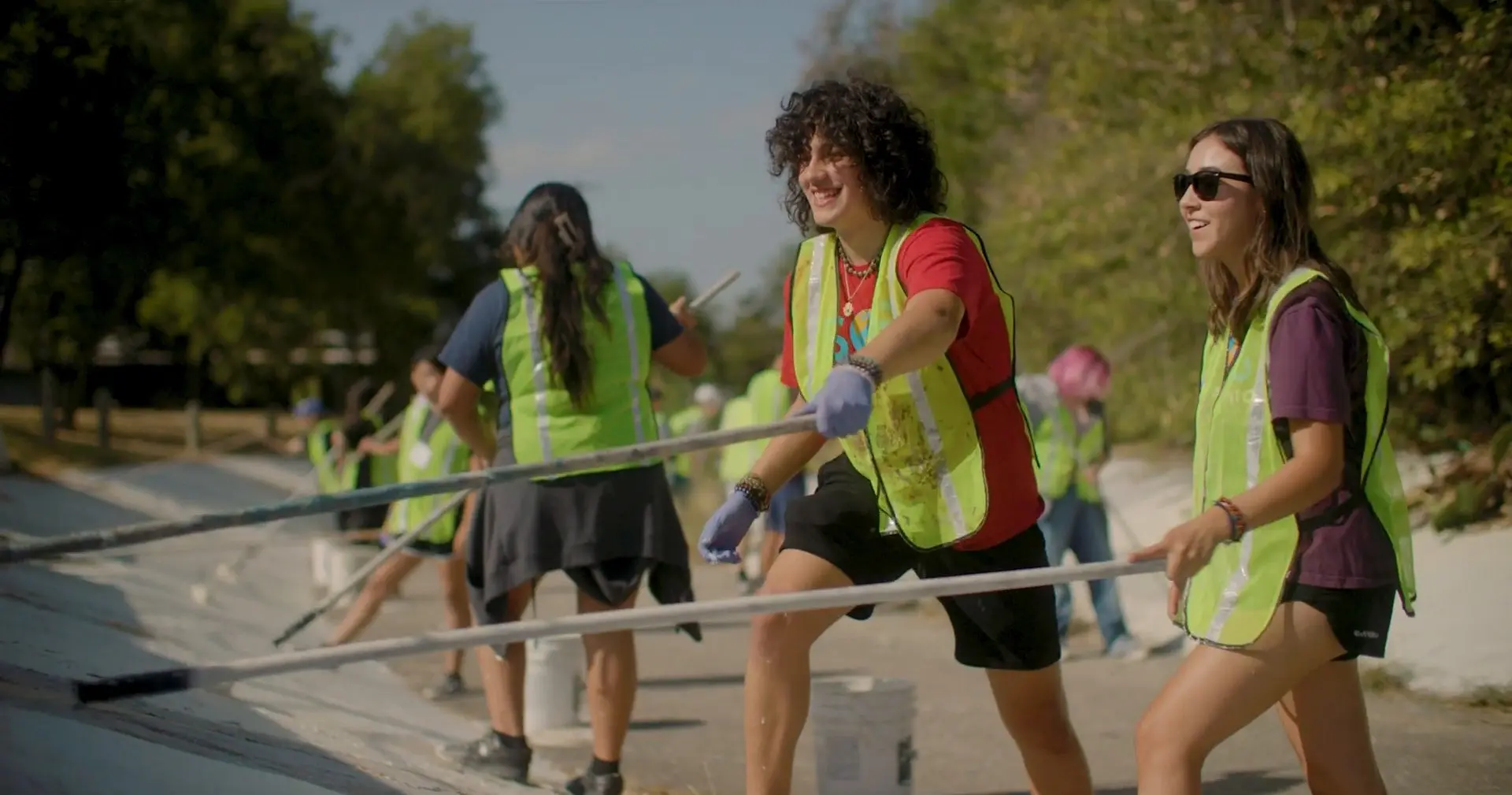 Smiling teen volunteers in neon yellow safety vests push long-handled paint rollers along a sloped concrete wall.